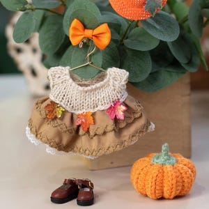May include: A miniature dress on a gold hanger with a cream knit bodice and a tan tiered skirt featuring decorative stitching and three small colorful leaf appliques. An orange bow is attached to the hanger, with brown shoes and a crocheted pumpkin.