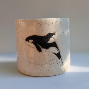 May include: A speckled ceramic mug with a black and white orca whale design. The orca is depicted in a stylized, minimalist form, adding a touch of marine life to the mug's aesthetic. The mug has a cylindrical shape.