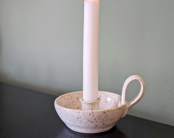 May include: A white ceramic candle holder with a speckled finish and a single handle. A white candle is burning in the holder.