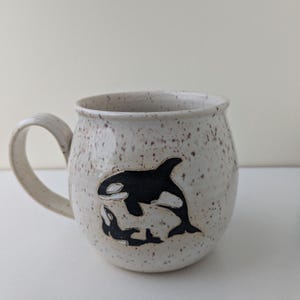 May include: A white ceramic mug with a speckled glaze features a black and white silhouette of an orca and calf.