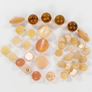 May include: An assortment of vintage beads and buttons in various shapes and sizes. The collection includes amber-colored, round beads, textured yellow beads, and flat, circular buttons in shades of peach and cream. Two wooden barrel-shaped beads are also present.