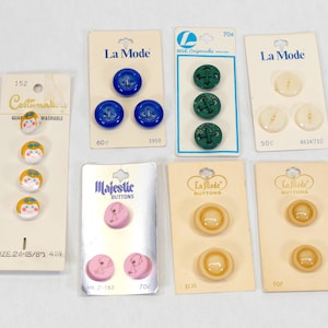 May include: A collection of vintage buttons on their original cards. The buttons vary in color and design, including blue, green, and cream-colored buttons. Some cards feature the brand names "La Mode" and "Majestic Buttons."