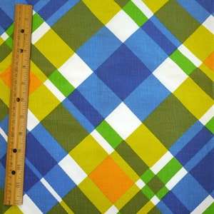 May include: A colorful fabric with a plaid pattern in shades of blue, green, yellow, white, and orange. A wooden ruler is placed vertically along the left side, showing inch measurements.