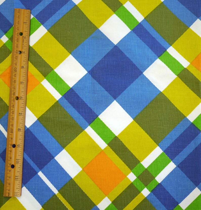 May include: A colorful fabric with a plaid pattern in shades of blue, green, yellow, white, and orange. A wooden ruler is placed vertically along the left side, showing inch measurements.