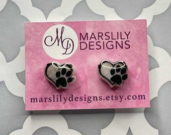 May include: A pair of heart-shaped stud earrings with a paw print design. The earrings are black and white and are displayed on a pink card with the text "MARSLILY DESIGNS" and the website marslilydesigns.etsy.com.