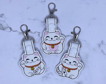 May include: Three white lucky cat keychains with embroidered details. Each cat has a pink outline, a smiling face, and a gold bell collar. The keychains have silver-colored clasps and white leather straps.