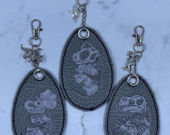 May include: Three gray dinosaur-themed keychains. Each keychain features a different dinosaur skeleton design, with a silver-colored charm of a dinosaur attached. The keychains are oval-shaped and have a textured appearance.