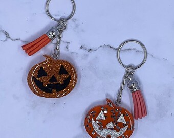 May include: Two Halloween-themed keychains. Each keychain features a glittery orange pumpkin charm with a carved face. The charms are attached to a silver chain and ring, with an orange tassel. The background is a light, marbled surface.