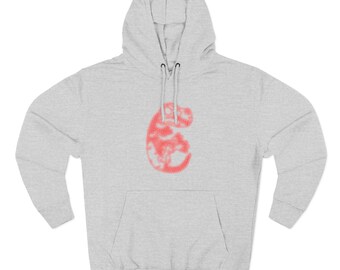 May include: A light gray hoodie with a red, abstract design on the front. The hoodie has a drawstring hood and a kangaroo pocket. The design appears to be a stylized shape with a textured pattern.