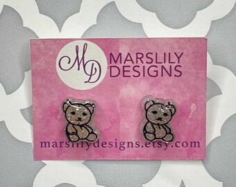 May include: A pair of teddy bear stud earrings on a pink card. The earrings are clear with a glittery fill and black outlines. The card has the text "Marslily Designs" and the shop's Etsy URL.