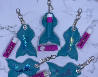 May include: A collection of teal mermaid tail-shaped lip balm holders with silver keychains. Each holder features a holographic mermaid design. Several lip balms in pink tubes are also visible, with labels reading "Vanilla Lavender" and "Sweet Orange/Lemongrass."