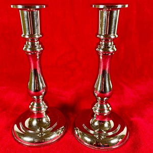 May include: Two silver-colored candlesticks with a reflective finish. Each candlestick has a wide, rounded base, a slender, curved stem, and a flat top. The candlesticks are displayed against a red background.