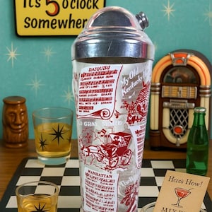Puede incluir: Una coctelera vintage con tapa plateada y cuerpo de vidrio transparente, con recetas de c&oacute;cteles e ilustraciones impresas en rojo. La imagen incluye un letrero que dice "It's 5 o'clock somewhere" y una peque&ntilde;a tarjeta que dice "Mixer for the Host".