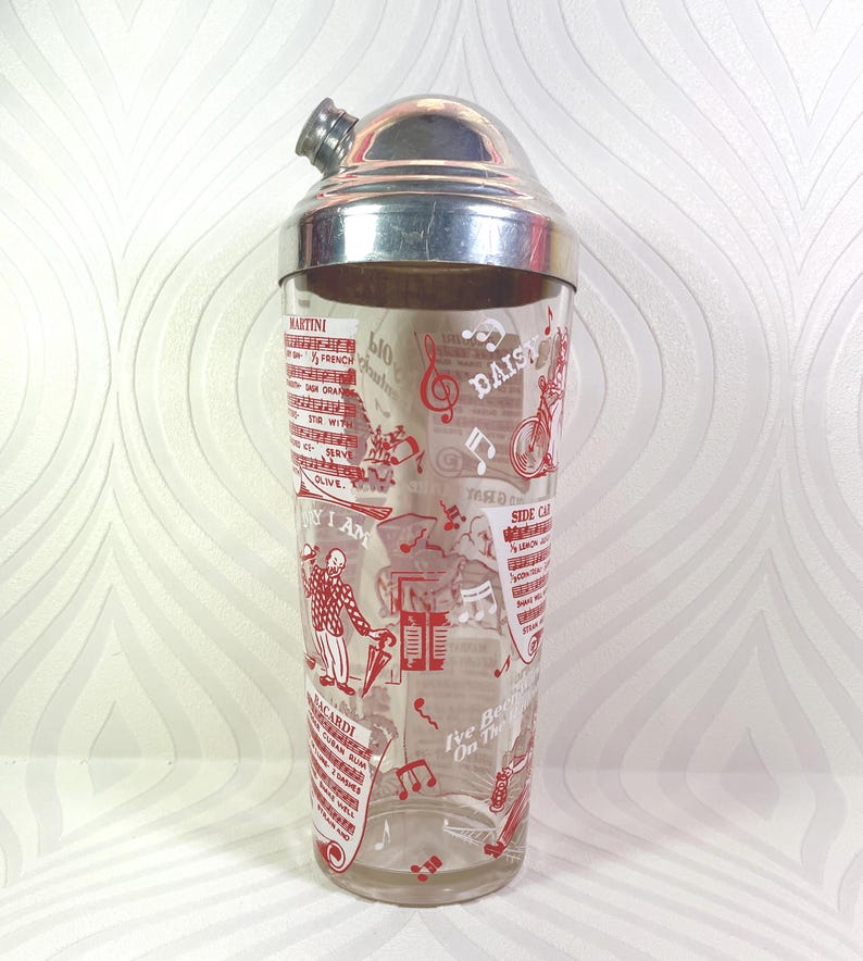 Puede incluir: Una coctelera vintage con tapa plateada y cuerpo de vidrio transparente. El vidrio est&aacute; decorado con ilustraciones rojas de notas musicales, recetas de c&oacute;cteles y las palabras "MARTINI", "DAISY" y "BACARDI".