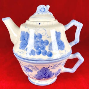 May include: A white ceramic teapot and teacup set with blue grape and leaf designs. The teapot has a lid with a floral accent and a spout. The teacup is stacked beneath the teapot, with a handle on each piece.