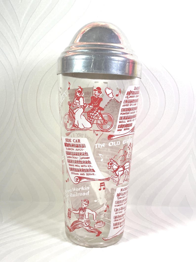 Puede incluir: Coctelera vintage con tapa plateada y cuerpo de vidrio transparente. El vidrio est&aacute; decorado con ilustraciones rojas de personas, notas musicales y recetas de c&oacute;cteles. El texto incluye "Side Car" y "The Old Grand".