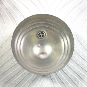 Puede incluir: Vista superior de un cuenco de metal plateado. El cuenco tiene forma circular con anillos conc&eacute;ntricos y un peque&ntilde;o emblema decorativo en el centro. La superficie met&aacute;lica refleja la luz, resaltando su textura.