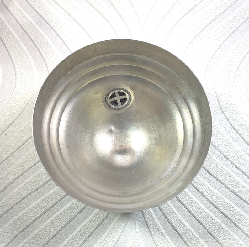 Puede incluir: Vista superior de un cuenco de metal plateado. El cuenco tiene forma circular con anillos conc&eacute;ntricos y un peque&ntilde;o emblema decorativo en el centro. La superficie met&aacute;lica refleja la luz, resaltando su textura.