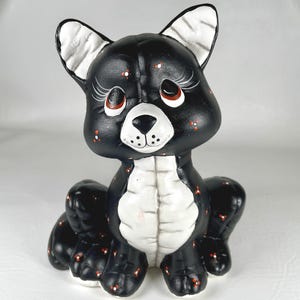 May include: A black and white ceramic cat figurine featuring large eyes with long painted eyelashes, a white muzzle, and a white chest. The black areas are decorated with small orange and white dot patterns, and the ears have white interiors.