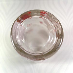 Puede incluir: Un cenicero de vidrio transparente con forma redondeada. El borde del cenicero tiene dise&ntilde;os florales rojos. El cenicero est&aacute; vac&iacute;o y se encuentra sobre una superficie blanca con un patr&oacute;n sutil.