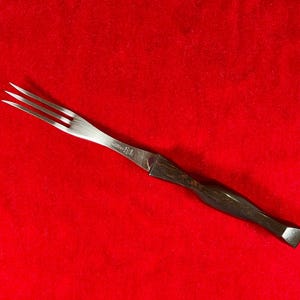 May include: A stainless steel fork with a dark wooden handle. The fork has three tines and a sharp blade. The handle is curved and has a geometric end. The fork is on a red background.