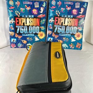 May include: Two hardcover books titled "Art Explosion 750,000 Images" with colorful illustrations. A gray, yellow, and black Case Logic CD case is in front of the books. The books are labeled "Volume One" and "Volume Two".