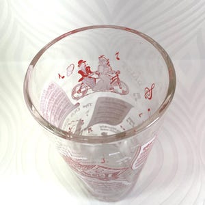 Puede incluir: Vaso de vidrio transparente con ilustraciones rojas. El dise&ntilde;o incluye una pareja en bicicleta t&aacute;ndem, notas musicales y texto. El vaso tiene una abertura ancha y un cuerpo ligeramente c&oacute;nico, adecuado para bebidas.