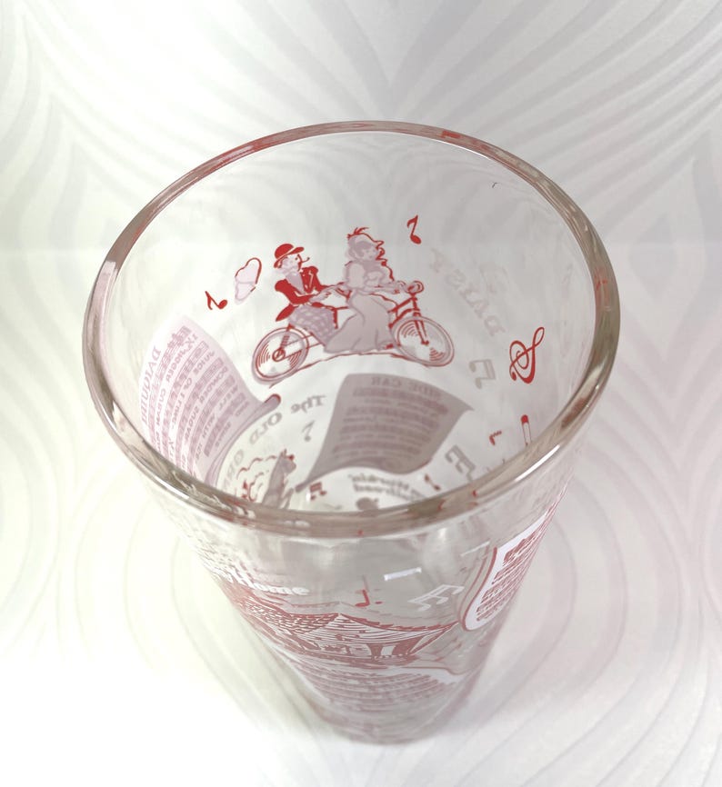 Puede incluir: Vaso de vidrio transparente con ilustraciones rojas. El dise&ntilde;o incluye una pareja en bicicleta t&aacute;ndem, notas musicales y texto. El vaso tiene una abertura ancha y un cuerpo ligeramente c&oacute;nico, adecuado para bebidas.