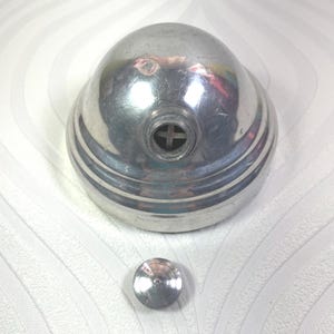 Puede incluir: Un objeto met&aacute;lico plateado vintage con una parte superior redondeada en forma de c&uacute;pula y una base circular. El objeto tiene una pieza circular peque&ntilde;a y separada. La parte superior tiene una abertura en forma de cruz. Probablemente sea un art&iacute;culo decorativo o funcional.