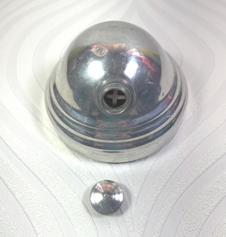 Puede incluir: Un objeto met&aacute;lico plateado vintage con una parte superior redondeada en forma de c&uacute;pula y una base circular. El objeto tiene una pieza circular peque&ntilde;a y separada. La parte superior tiene una abertura en forma de cruz. Probablemente sea un art&iacute;culo decorativo o funcional.