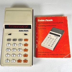 May include: A beige Radio Shack EC-475 scientific calculator with a red digital display and white and orange buttons, accompanied by a red operating instructions manual featuring the text Radio Shack EC-475 Scientific Calculator Cat. No. 65-634.