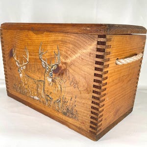 May include: A rectangular wooden storage chest featuring a wood-burned illustration of two deer standing in a forest setting. The box has finger-jointed corners and a thick rope handle attached to the side, with a lid covering the top opening.