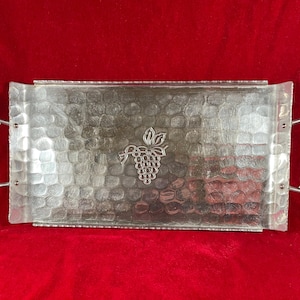 May include: Silver-toned rectangular serving tray with a textured surface and a grape design in the center. The tray has twisted metal handles on each end. The tray is displayed on a red background.