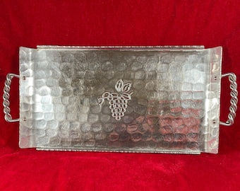 May include: Silver-toned rectangular serving tray with a textured surface and a grape design in the center. The tray has twisted metal handles on each end. The tray is displayed on a red background.