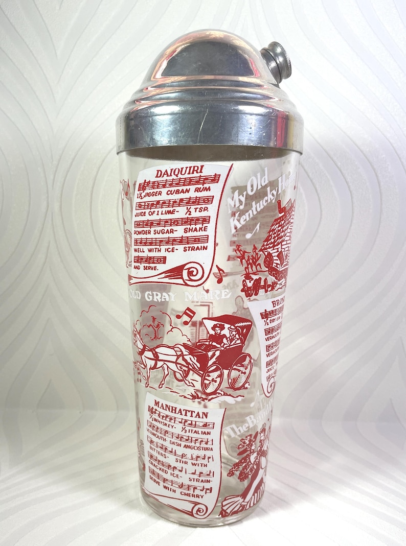 Puede incluir: Coctelera vintage con tapa de acero inoxidable y cuerpo de vidrio transparente. El vidrio est&aacute; decorado con ilustraciones y texto en rojo, incluidas recetas de c&oacute;cteles como Daiquiri y Manhattan.