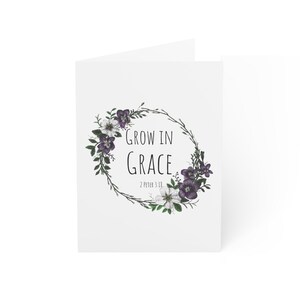 May include: A white greeting card featuring a circular wreath of purple and white flowers with green leaves. Centered inside the wreath is the black text "GROW IN GRACE" followed by the smaller text "2 PETER 3:18" in a simple sans-serif font.