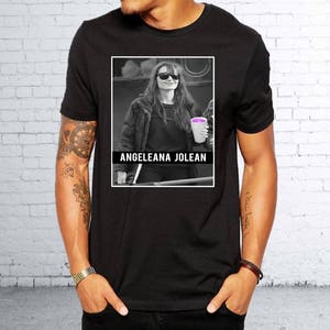 Könnte beinhalten: Schwarzes T-Shirt mit einem Foto einer Frau in einer schwarzen Jacke und Sonnenbrille, die ein lila Getränk in einem weißen Becher hält. Der Text "ANGELEANA JOLEAN" ist unter dem Foto gedruckt.