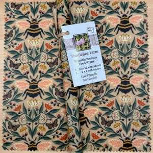 May include: Beeswax food wraps with a repeating pattern of bees, butterflies, and flowers in shades of beige, green, orange, and black. One wrap is rolled up. The label reads "ThistleBee Farm Reusable Beeswax Food Wraps".