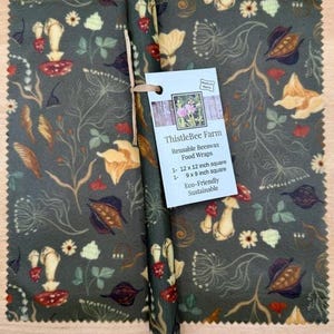 May include: Reusable beeswax food wraps with a dark green botanical print featuring mushrooms, flowers, and leaves. The wraps are 12 x 12 inches and 9 x 9 inches. The tag reads "ThistleBee Farm Reusable Beeswax Food Wraps Eco-Friendly Sustainable."