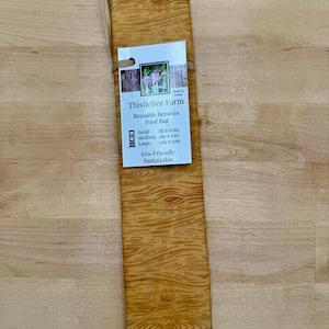 May include: A rectangular, reusable beeswax food bag with a wood grain pattern. The bag is labeled "ThistleBee Farm" and includes size options: Small (7in x 9.5in), Medium (9in x 13in), and Large (12in x 17in). The tag also states "Eco-Friendly Sustainable."
