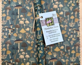 May include: Reusable beeswax food wraps with a mushroom pattern in shades of teal, brown, and yellow. The set includes one 12 x 12 inch square and one 9 x 9 inch square. The label reads "ThistleBee Farm Reusable Beeswax Food Wraps".