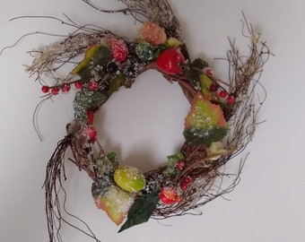 May include: A brown twig wreath decorated with red berries, green leaves, and faux fruit. The wreath is dusted with white glitter, giving it a snowy look.