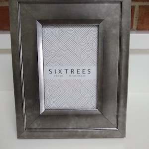 May include: A silver picture frame with a 4x6 inch opening. The frame has a simple, modern design and is made of metal. The frame is sitting on a brick surface.