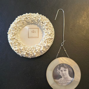 May include: Two white pearl picture frames. The larger frame is a round ornament with a hook for hanging. The smaller frame is a round tabletop frame with a white label that reads "sia Collection".