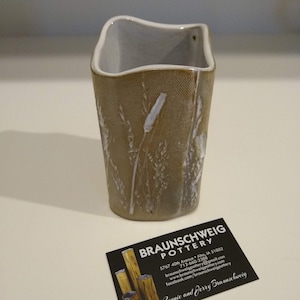 May include: A small, light brown ceramic vase with a white glaze and a textured surface. The vase has a square shape with rounded edges and features a design of white grass stalks.