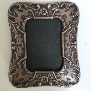 May include: Ornate copper-colored picture frame with a rectangular opening and a black backing. The frame features a detailed floral and scrollwork design. The frame is a decorative item for displaying photographs or artwork.