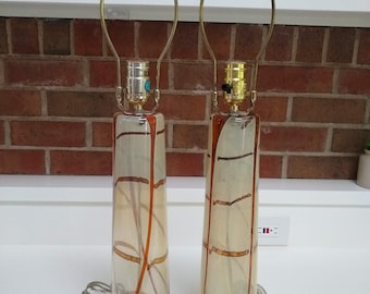May include: A pair of tall, cream-colored table lamps with gold accents and red glass finials. The lamps have a unique, textured glass design with a pattern of brown lines.