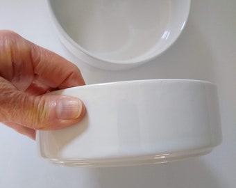 May include: Two white ceramic bowls, one larger than the other, with a smooth, glossy finish. The smaller bowl is being held in a hand.