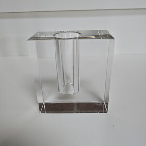 May include: A clear glass vase with a square base and a cylindrical opening. The vase is made of thick glass and has a polished finish.