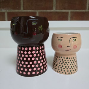 May include: Two ceramic planters with faces painted on them. The planter on the left is brown with pink polka dots and the planter on the right is light pink with green polka dots.